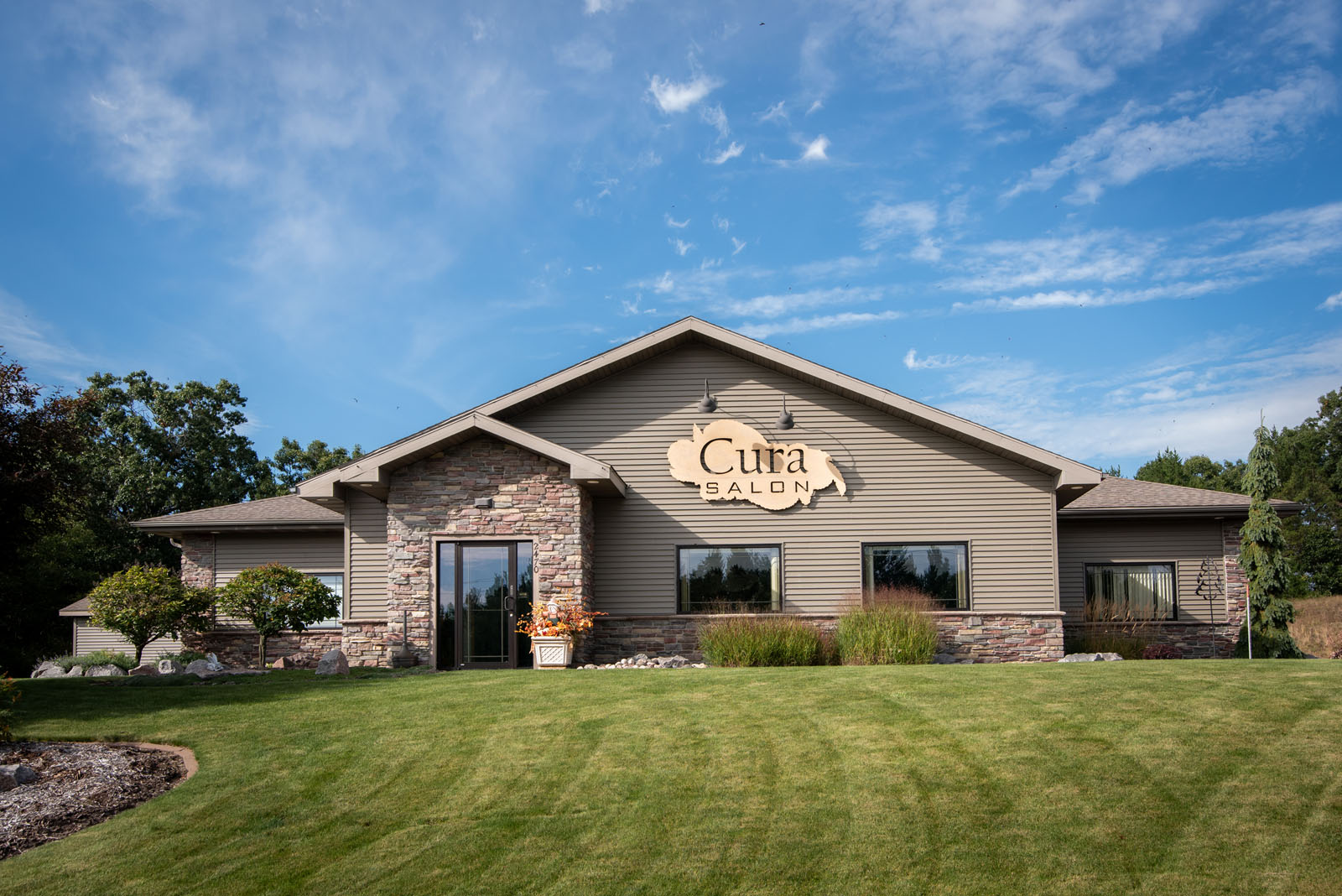 A single-story building with stone and tan siding, large windows, and a sign reading “Cura Salon.” The salon sits on a well-manicured lawn under a blue sky with scattered clouds.