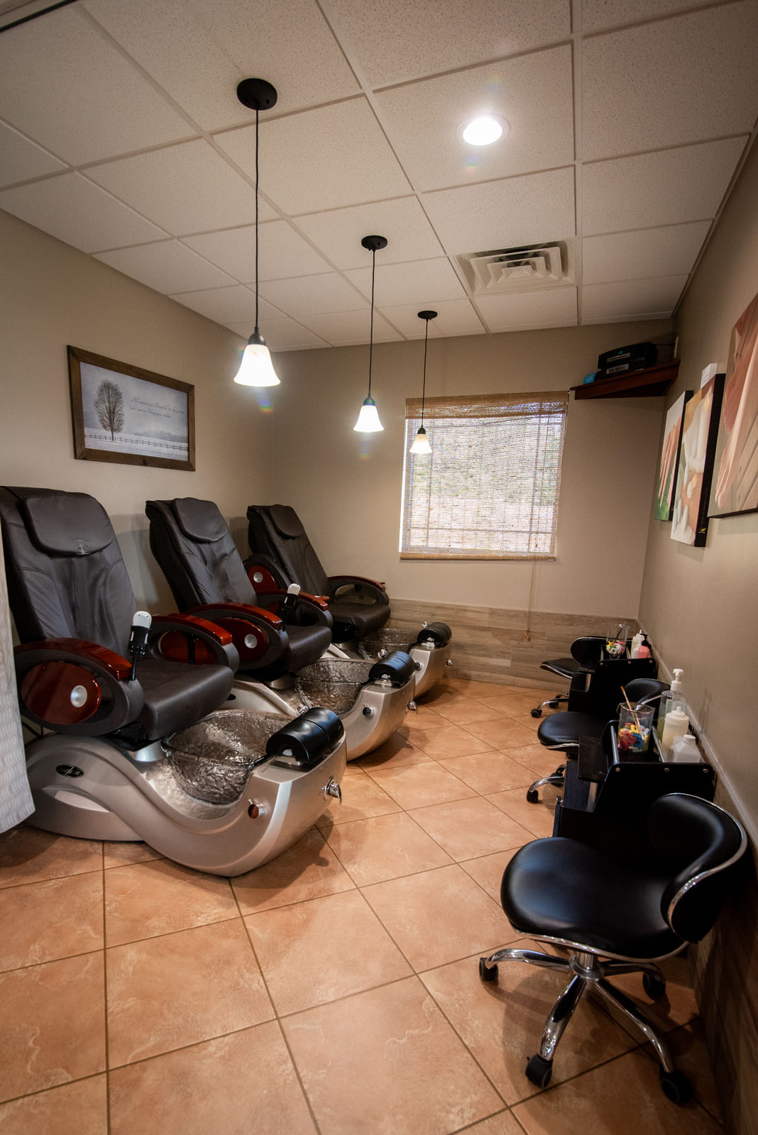 Three black pedicure spa chairs with foot baths are lined up in a tiled room. Two black stools and a small workstation with nail care supplies are on the right. Art and a window decorate the walls.