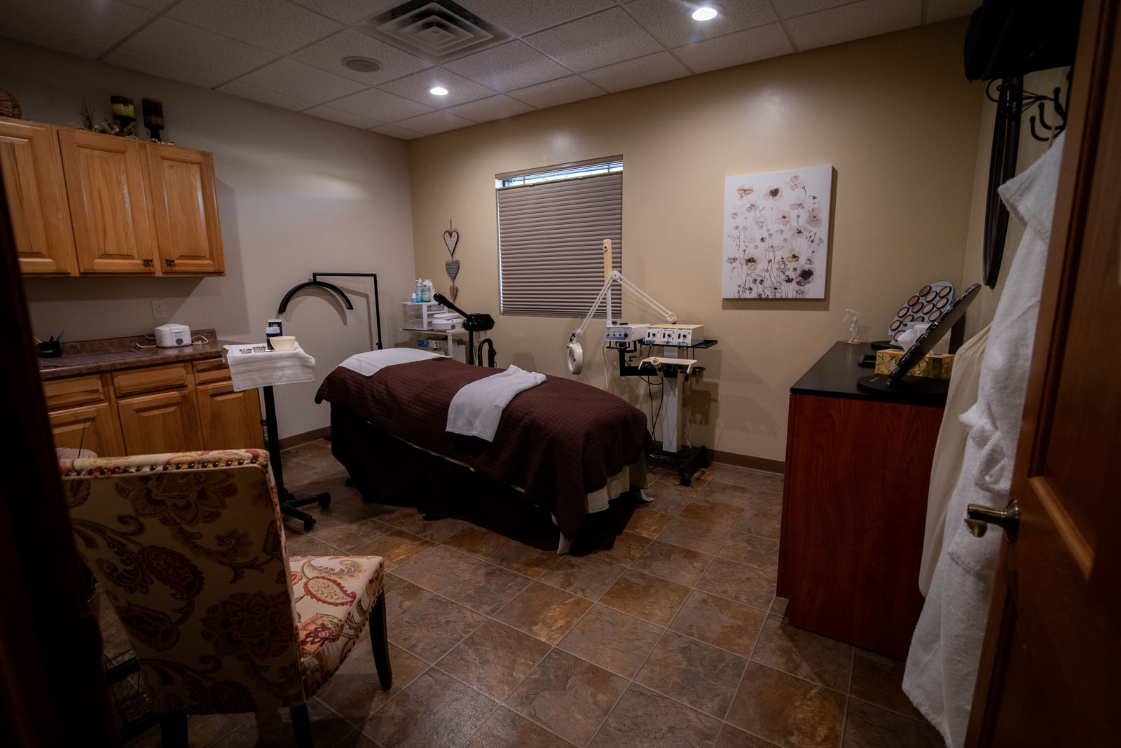 A spa treatment room with a massage table, cabinets, a decorative chair, various spa equipment, and soft lighting, creating a calm and relaxing atmosphere.