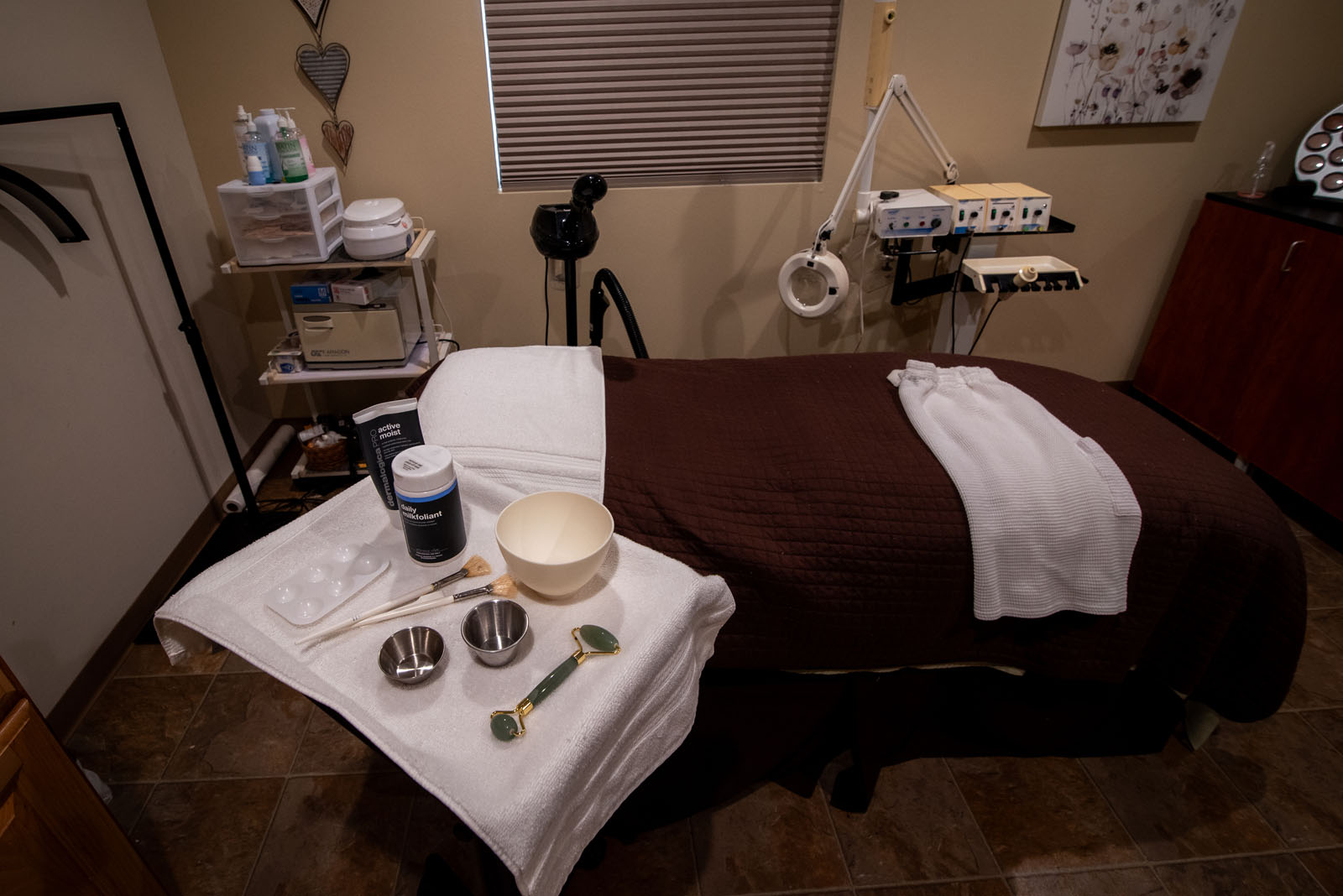 A spa treatment room with a massage table covered by brown and white towels. Nearby, bowls, a mixing spoon, and other spa tools are arranged on a small table. Shelves hold various bottles and equipment.