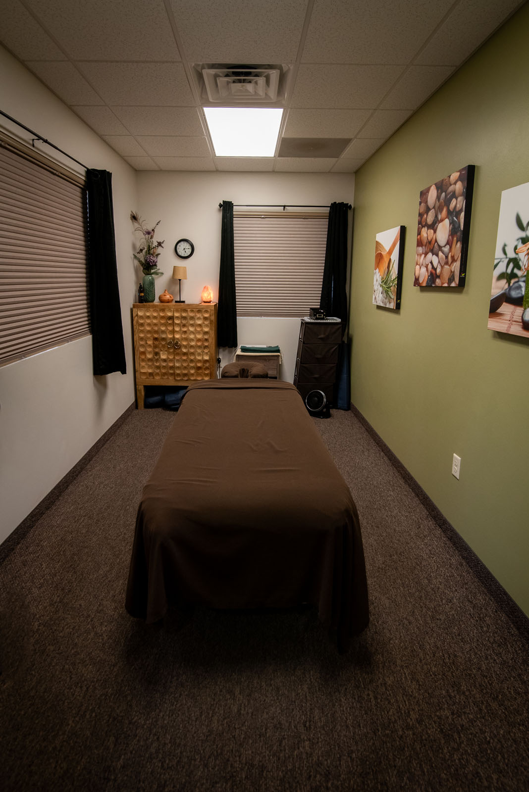 A massage therapy room with a bed covered in a brown sheet, soft lighting, blinds on windows, a wooden cabinet, and calming artwork on the green wall. The atmosphere is cozy and relaxing.