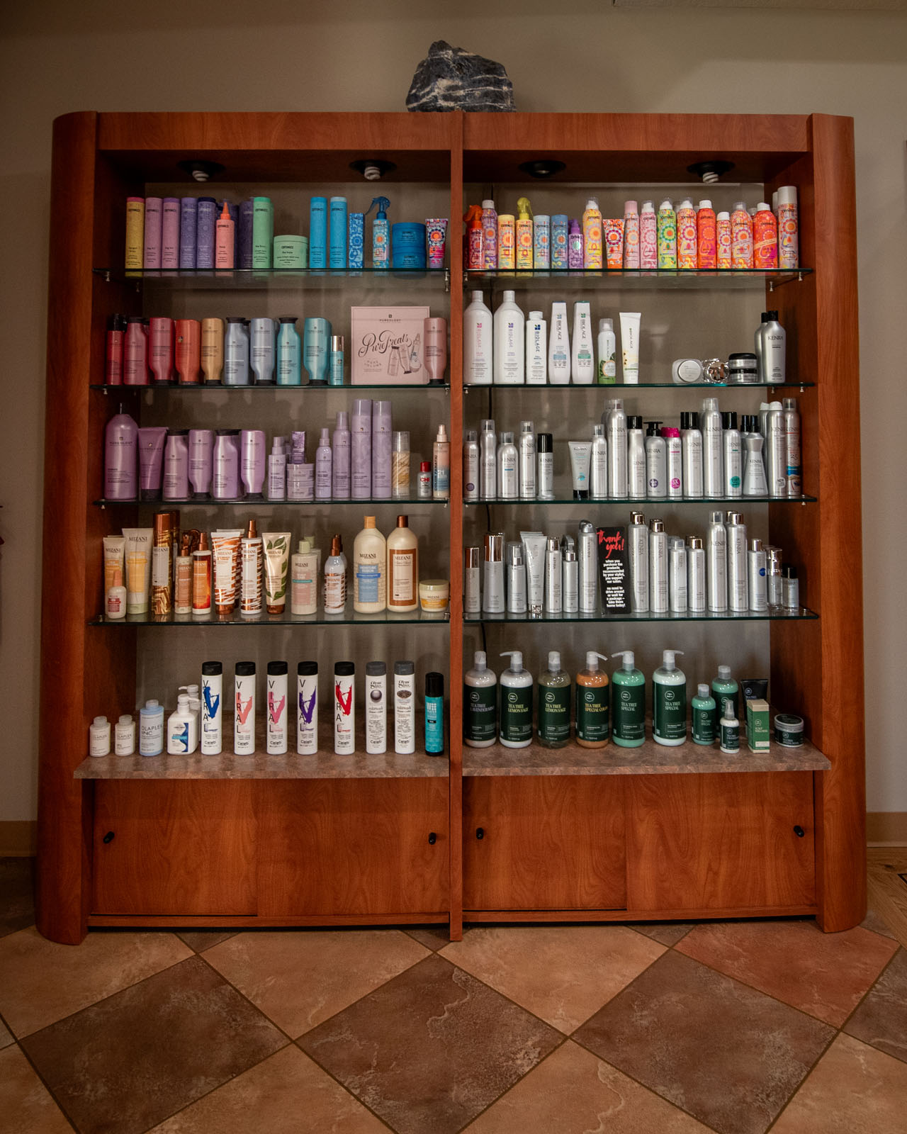 A wooden display shelf holds various colorful hair care products, including shampoos, conditioners, and styling sprays, arranged by type and brand on glass shelves against a tiled floor.