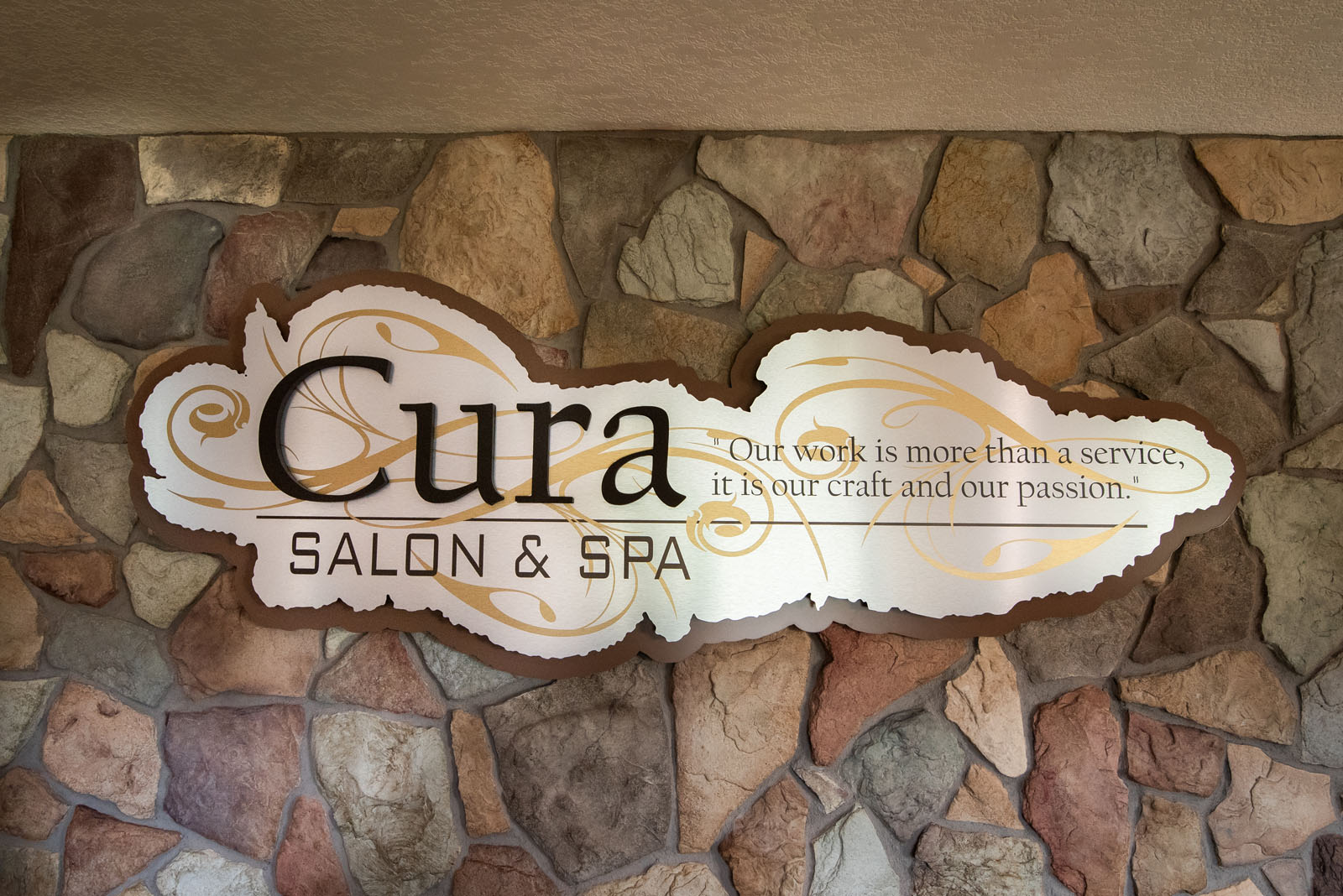 A sign reading “Cura Salon & Spa” with the quote “Our work is more than a service, it is our craft and our passion,“ mounted on a multicolored stone wall.