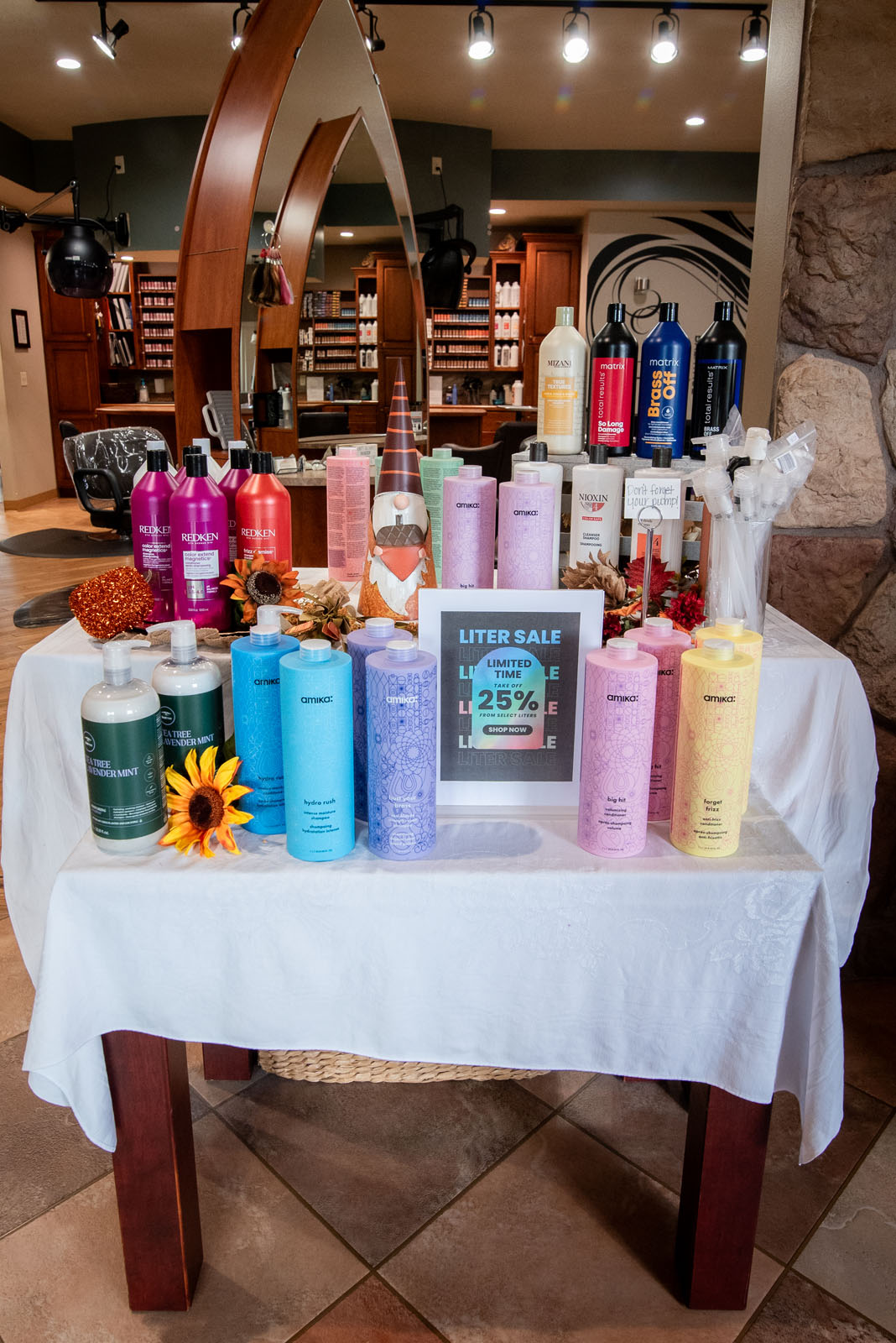 A display table in a salon features assorted shampoo and conditioner bottles, a “Liter Sale Up to 25% Off” sign, autumn leaves, and artificial sunflowers on a white tablecloth. Shelves of hair products are in the background.