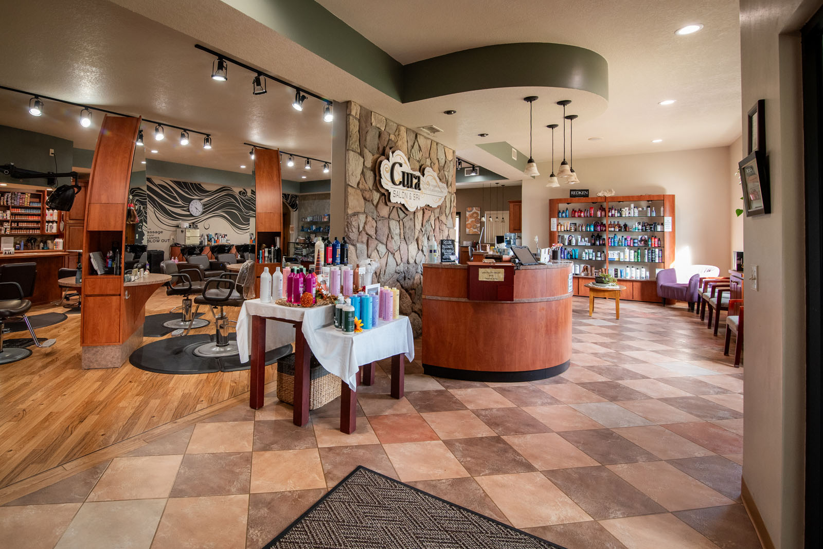 A modern salon interior with hairstyling stations, a reception desk, shelves of products, and a display table with bottles. The floor is tiled, and lighting is warm and inviting.