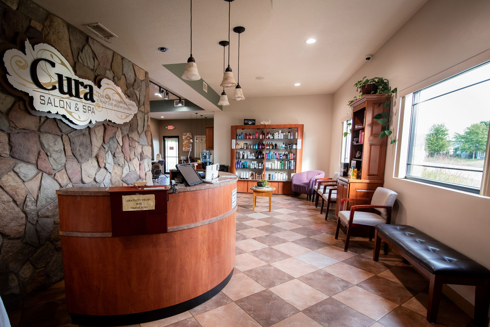 Reception area of a salon and spa with a curved desk, stone accent wall, product shelves, chairs, and large window overlooking greenery. The floor is tiled in a checkered pattern.
