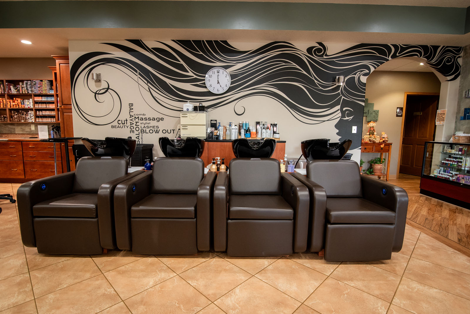 Four brown salon wash chairs are lined up in front of sinks. The wall behind features a black and white mural with flowing hair and a clock. Shelves with products are visible on the left and right.