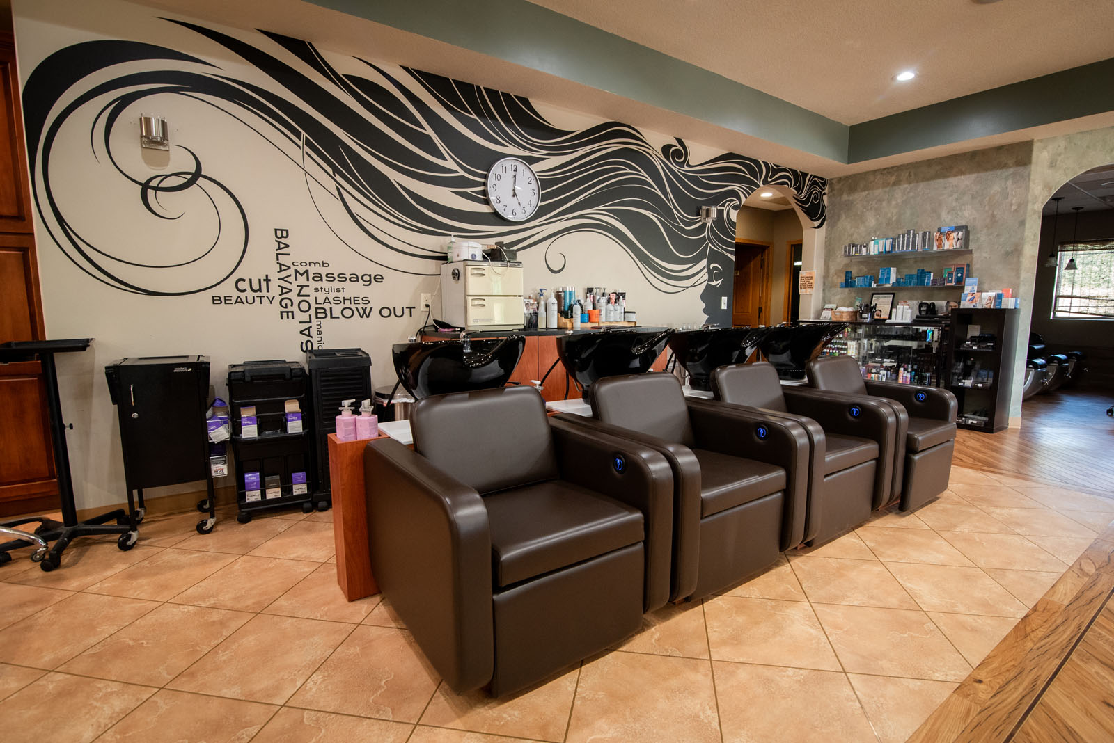 Three black salon chairs face washbasins in a modern hair salon with a decorative mural, shelves of hair products, and tiled flooring. A clock hangs above the chairs on the mural-adorned wall.