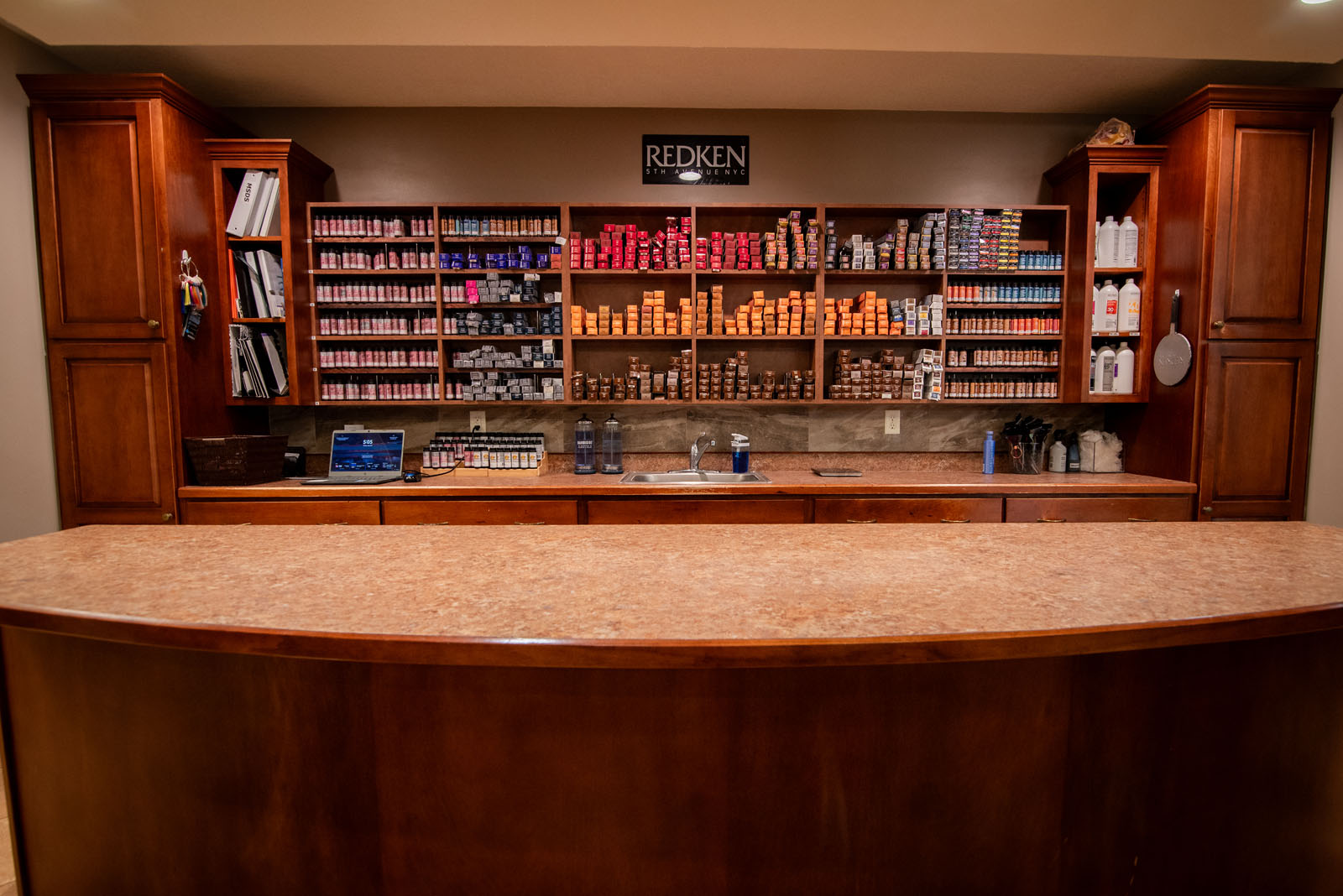 A salon counter with wooden cabinets and shelves filled with hair products, bottles, and hair color boxes. A laptop and supplies are on the counter; a “Redken” sign is above the shelves.