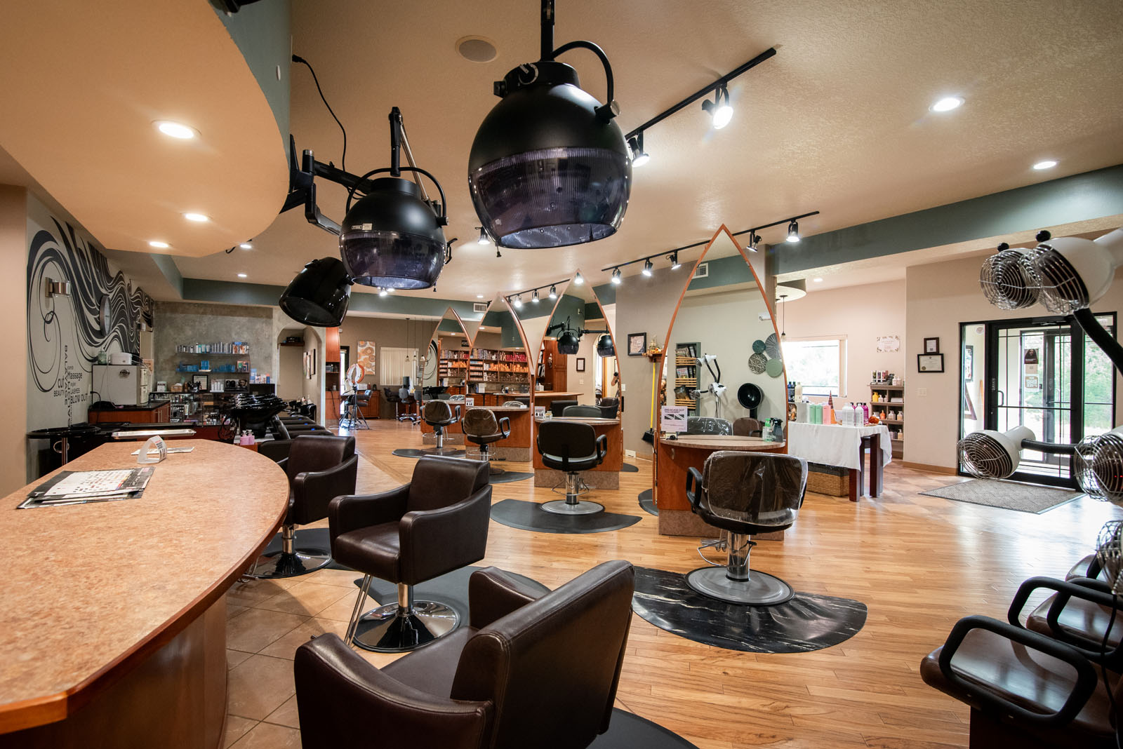 A modern hair salon interior with empty styling chairs, hair dryers, large mirrors, wooden floors, and shelves with hair products. The space is bright, clean, and spacious.
