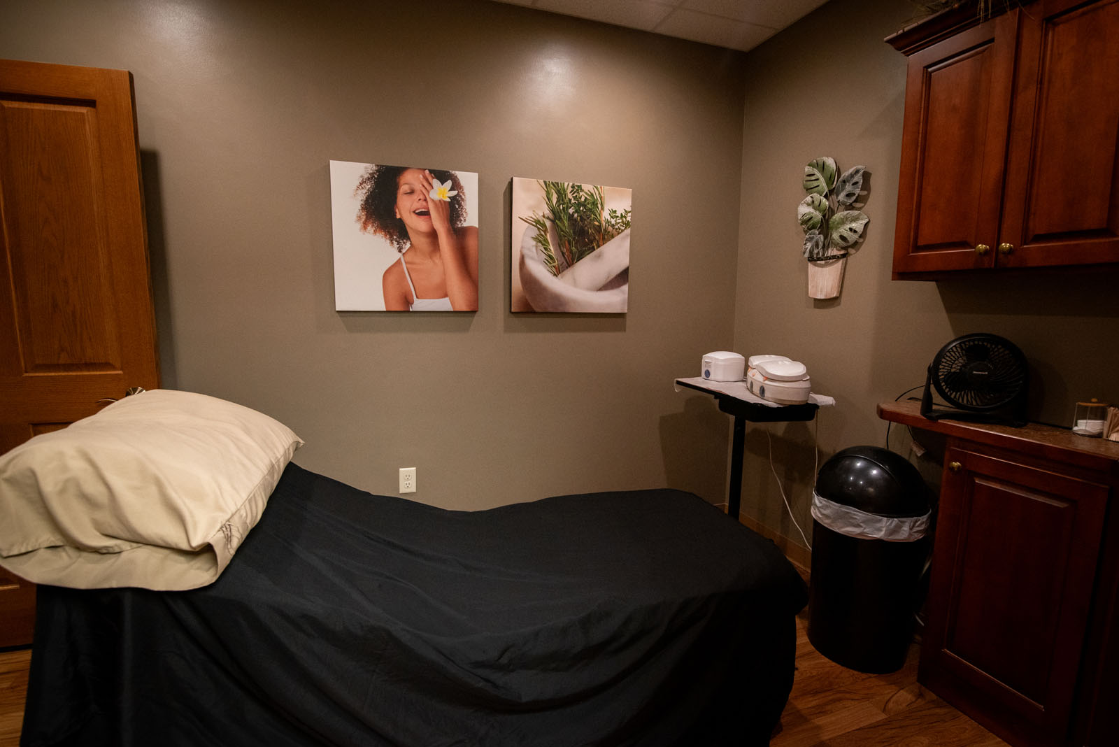 A cozy spa room with a black-covered massage table, soft lighting, wooden cabinets, artwork of a smiling woman and plants, and spa equipment on a small table.
