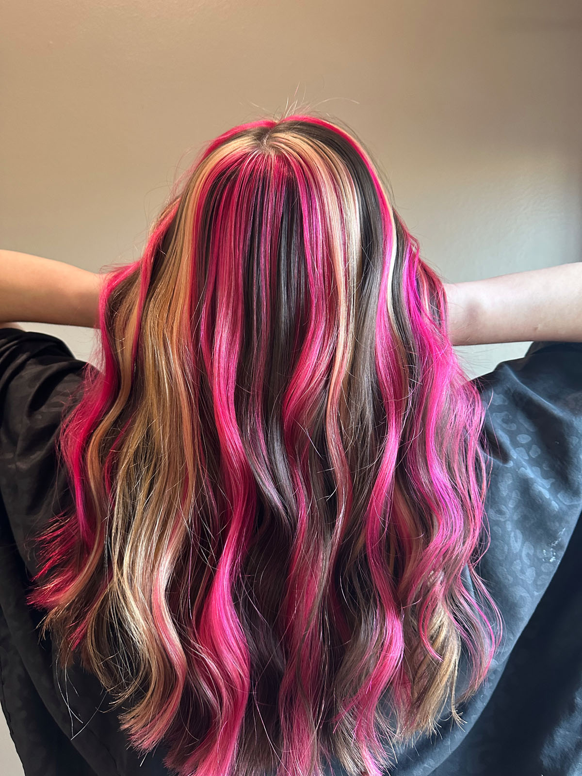 Person with wavy hair featuring vibrant pink, blonde, and brown highlights, viewed from behind against a plain background. Their arms are raised, elbows bent, and they wear a dark garment or cape.