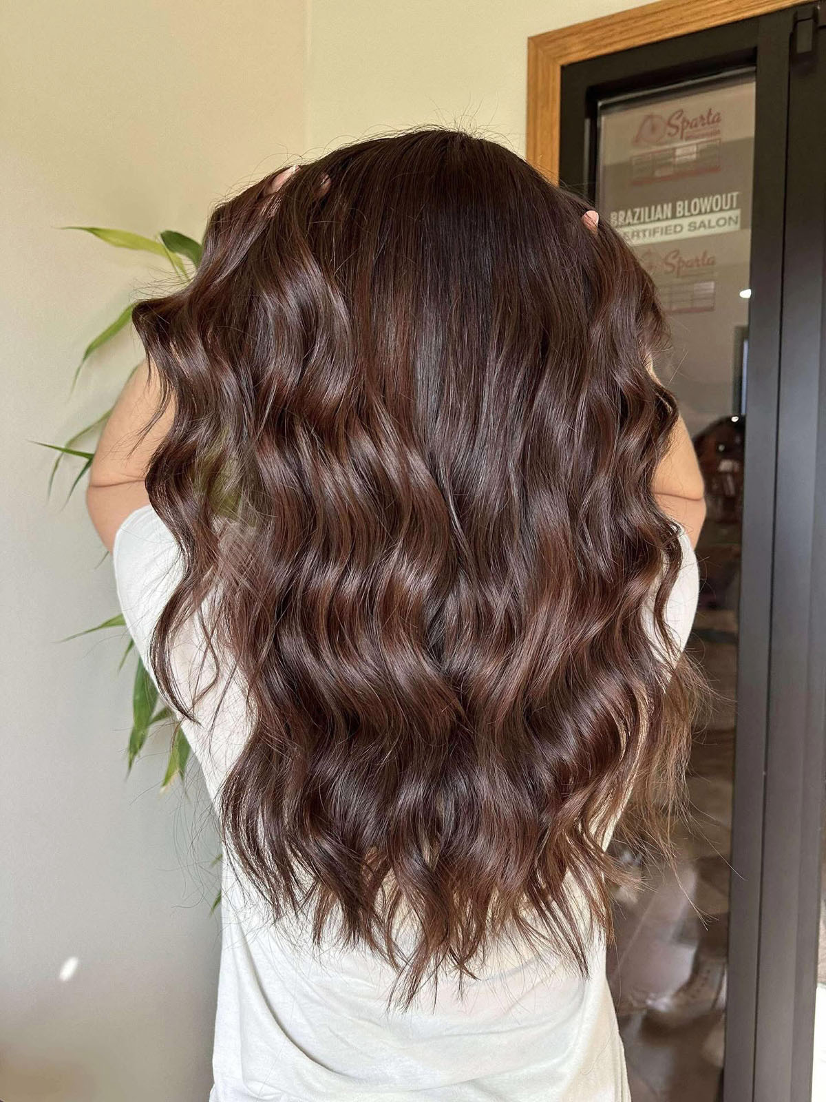 A person with long, wavy, brown hair stands facing away, hands in their hair, wearing a white shirt. A plant and a glass door with a sign are visible in the background.