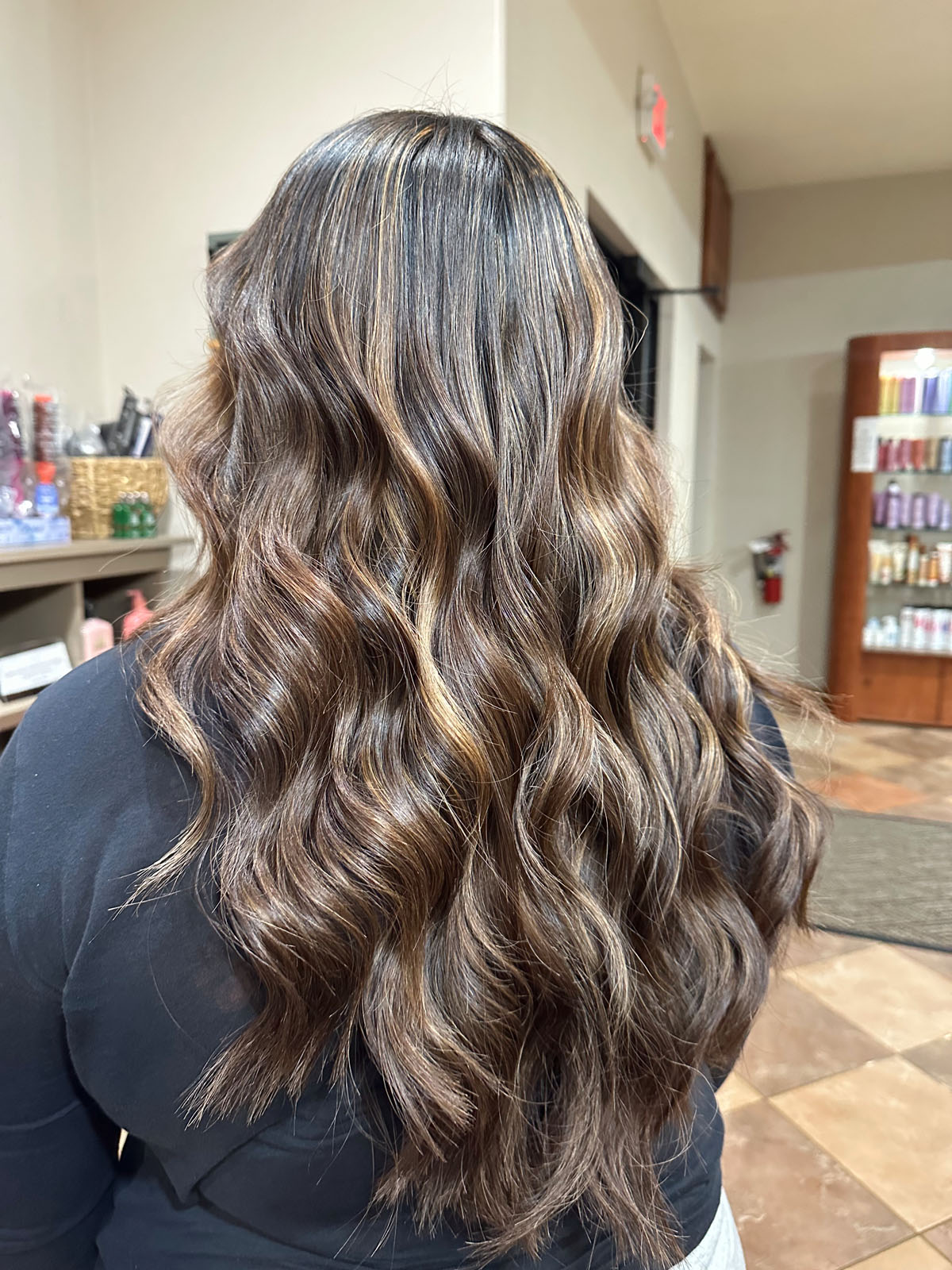 A person with long, wavy brown hair featuring caramel highlights stands indoors near salon shelves, facing away from the camera.