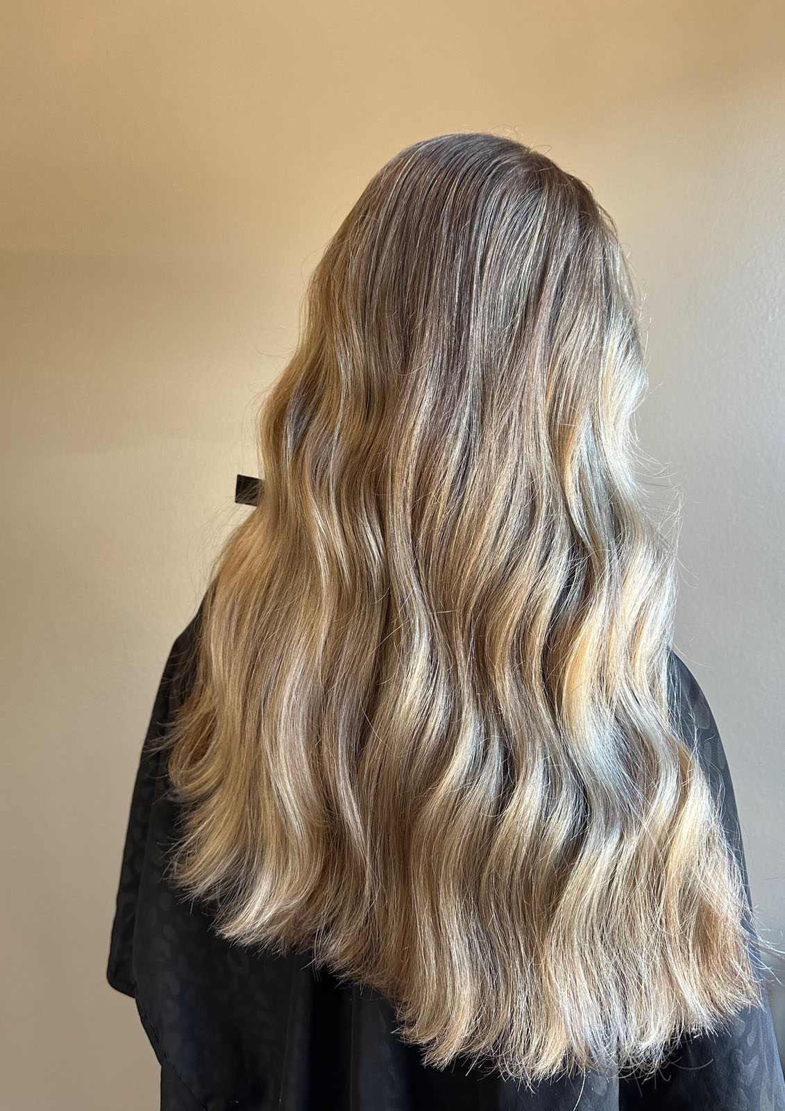 A person with long, wavy, blonde hair stands facing away from the camera, wearing a black salon cape against a plain beige background.