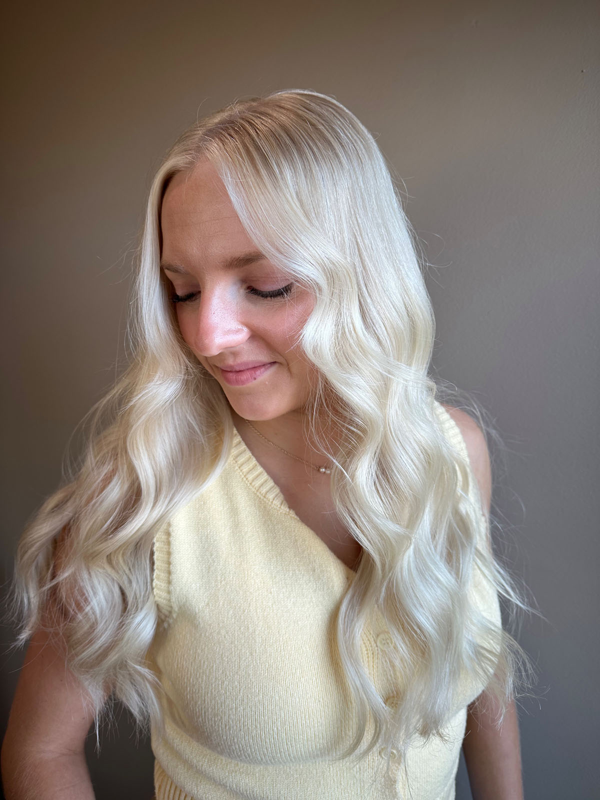 A person with long, wavy platinum blonde hair and a side part is wearing a light yellow sleeveless top, looking down and slightly smiling against a plain, neutral background.