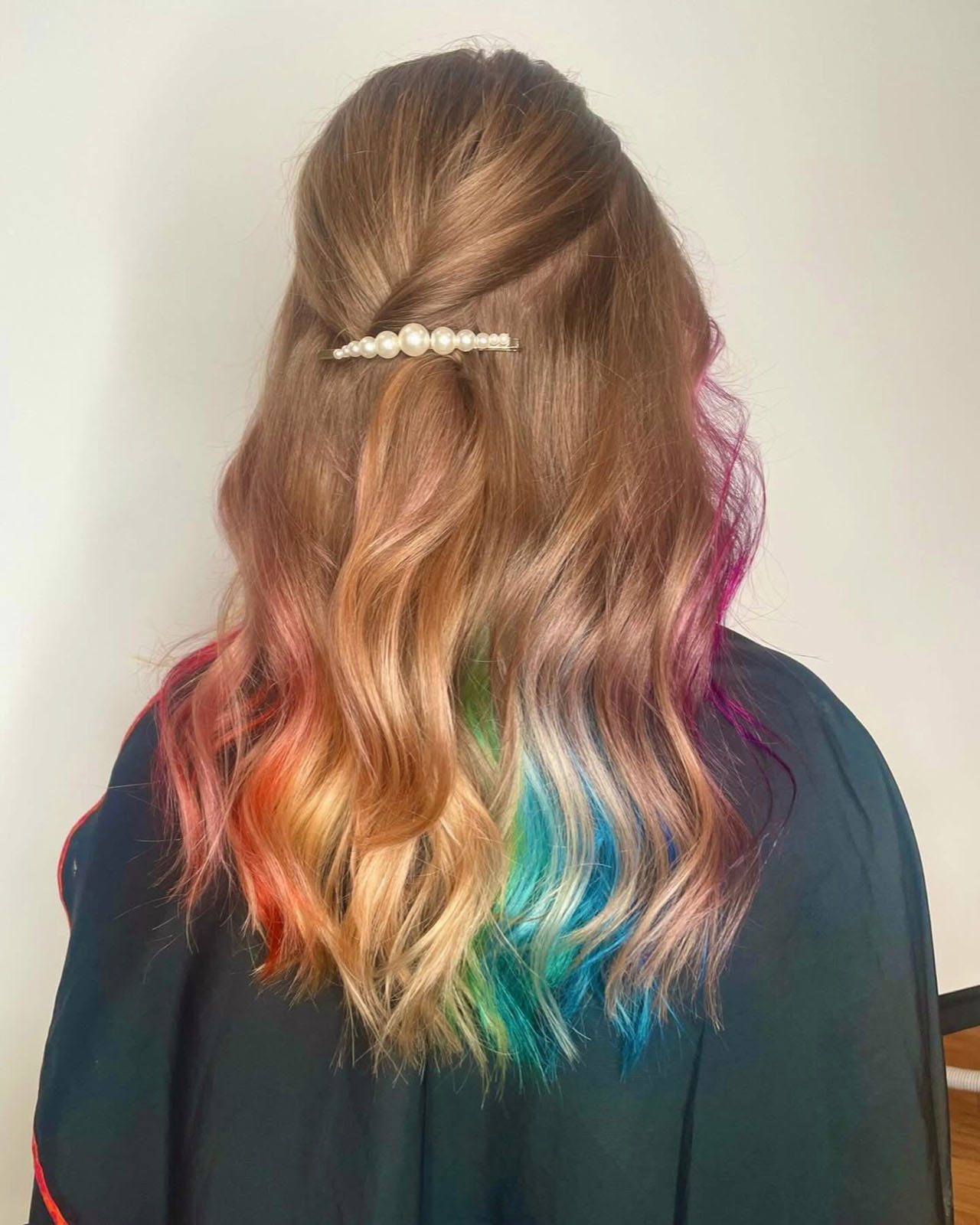 A person with wavy brown hair styled half-up, secured with a pearl hair clip. The hair ends are dyed in a rainbow of colors: red, orange, green, blue, and purple. The background is plain and light-colored.