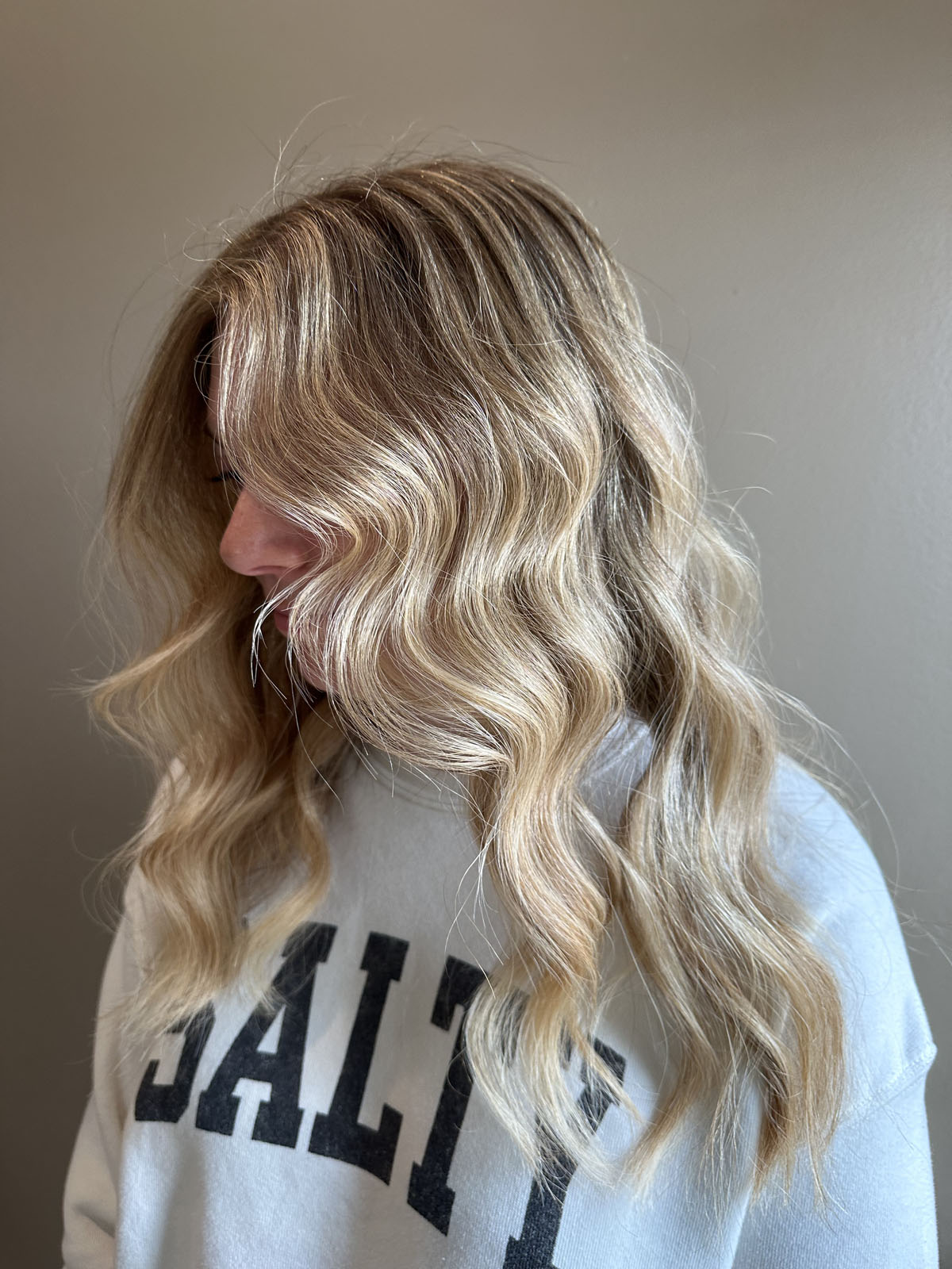 A person with long, wavy blonde hair is shown in profile, wearing a light-colored sweatshirt with the word “SALTY” in bold black letters. The background is plain and neutral.